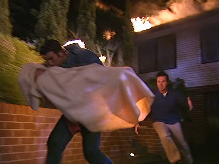 Screencap of Drew Kirk running out of the fire at 26 Ramsay Street carrying Louise Carpenter wrapped in a blanket. Drew has caught fire, with Karl Kennedy closely behind him.