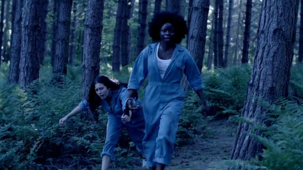 Lauren drags Mitzeee through a forest by the handcuffs they are joined with. Both are wearing prison attire.