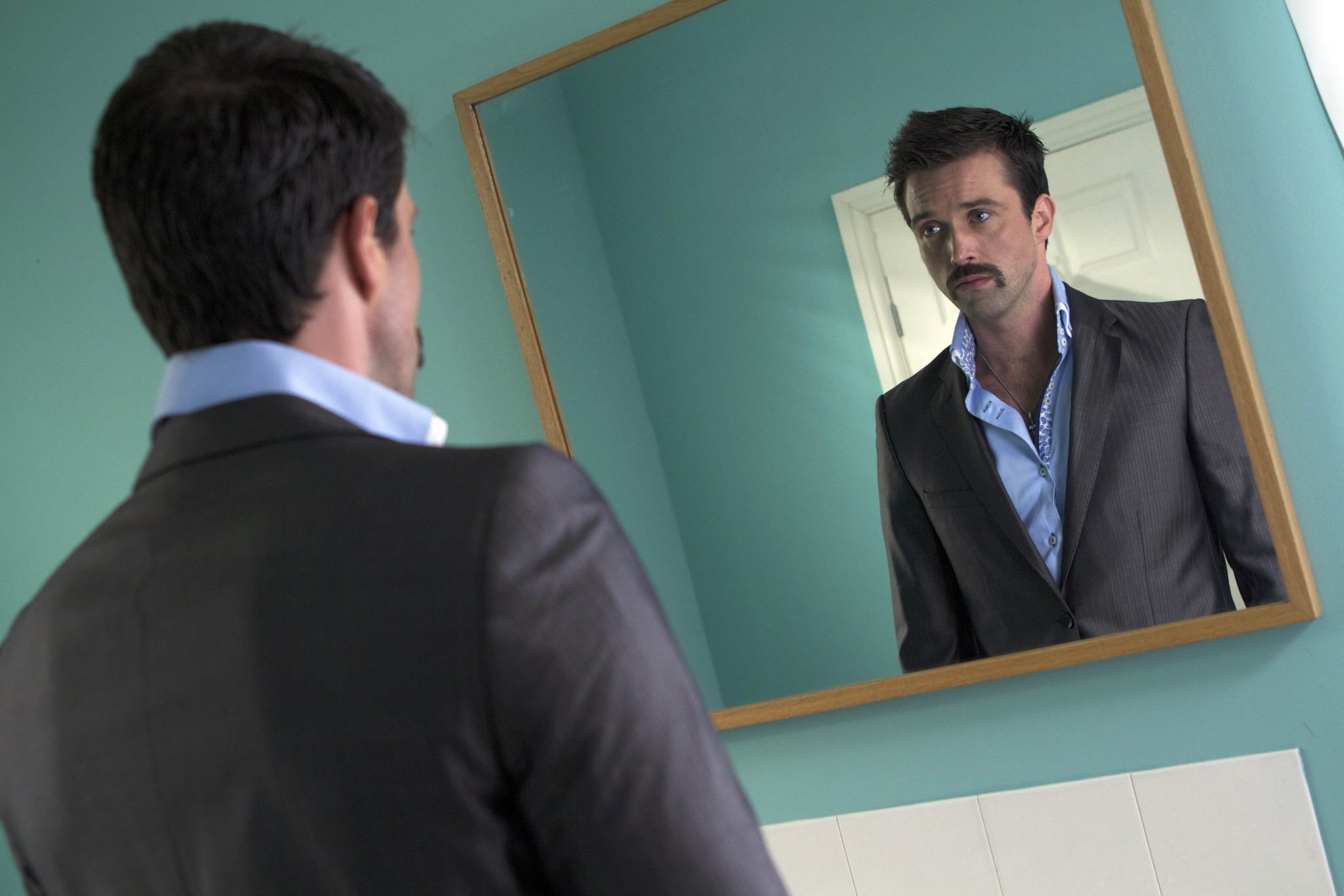 Brendan looks at his reflection in a mirror. He is wearing a smart suit and is well-groomed.