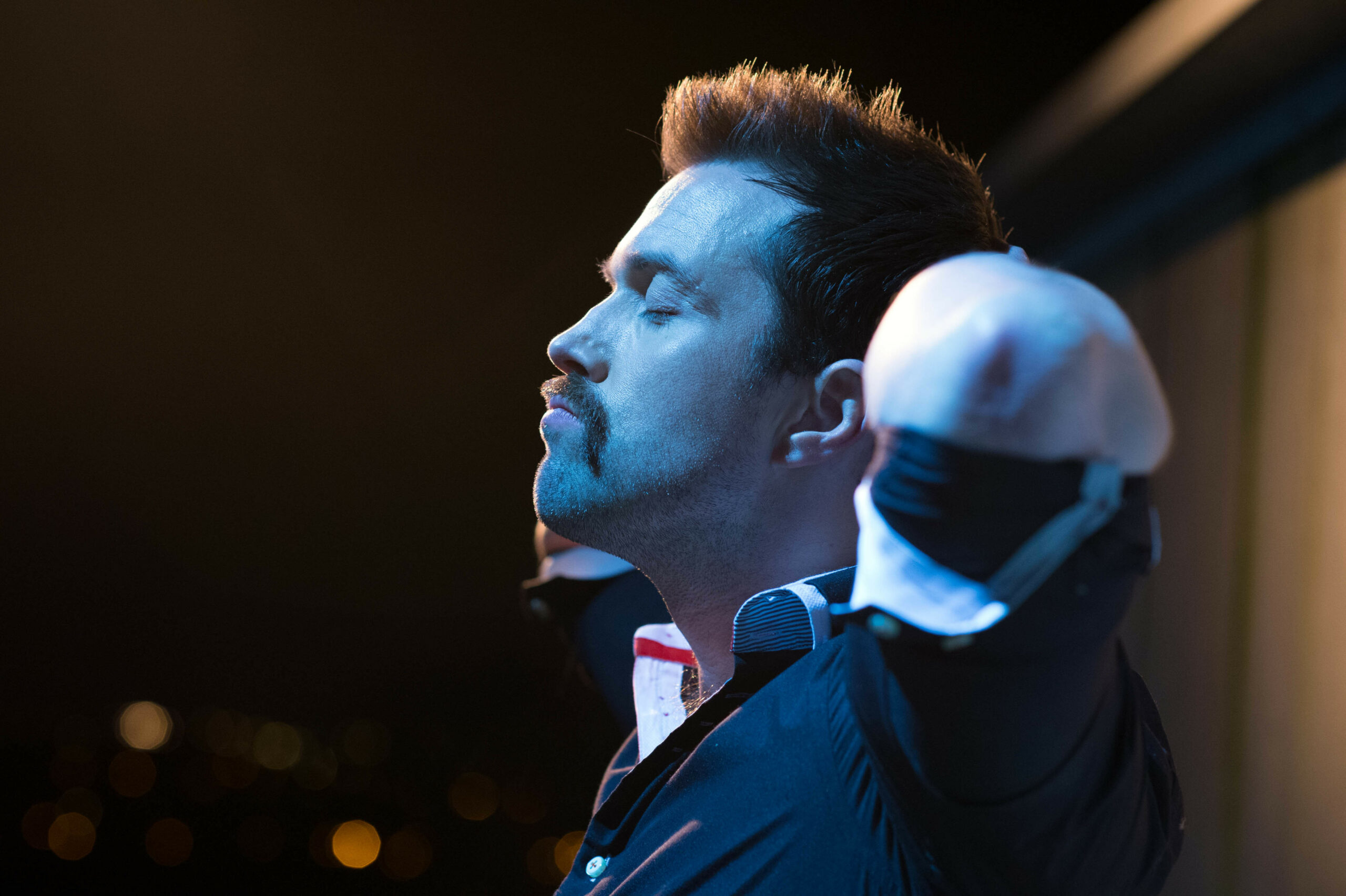 Brendan stands outside, lit with blue hue light, hands behind his head.