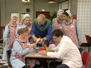 dolly, bren, jean, stan, twinkle, tony and anita sit around a table as stan shows bren a piece of paper